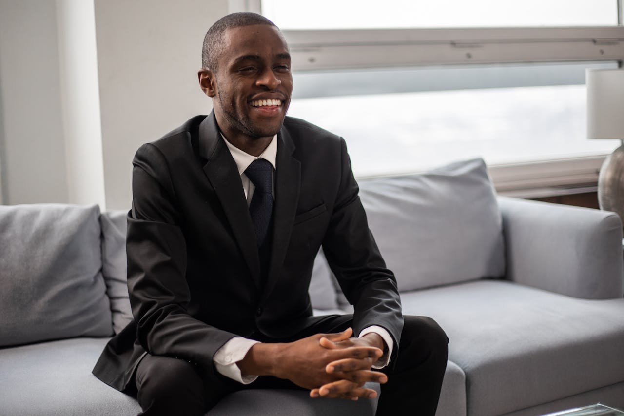 A smiling black man in formal wear sitting on a sofa indoors, exuding confidence.